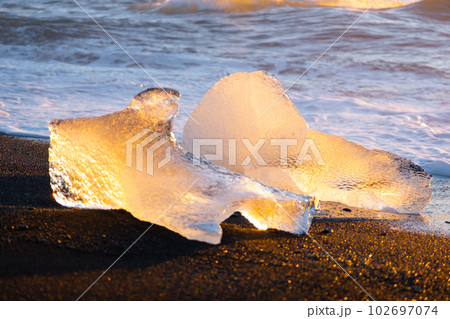 Pure glacier ice shining at sunset. Black volcanic sand at ocean coast. Pure ocean waves crash golden icebergs in Iceland. Diamond beach - famous tourist location in north Europe country. 102697074