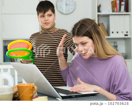 Mother working at laptop and unhappy son standing racquets for tennis 102703628