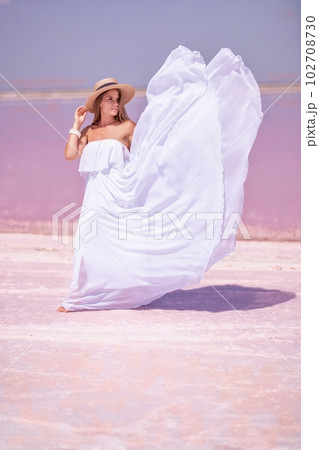 Woman in pink salt lake. She in a white dress and hat enjoys the scenic view of a pink salt lake as she walks along the white, salty shore, creating a lasting memory. 102708730