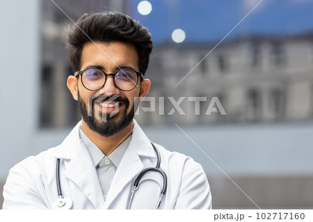 Close-up portrait of Hindu doctor, man in glasses smiling and looking at camera, doctor student intern with stethoscope outside clinic. Close-up portrait of Hindu doctor, man in glasses smiling and looking at camera, doctor student intern with stethoscope outside clinic. 102717160