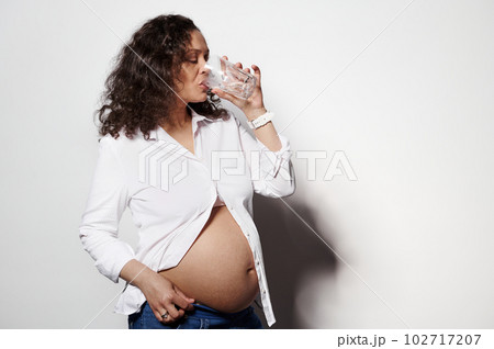Conscious multi ethnic charming adult pregnant woman drinking water and taking medicine or prenatal vitamins, posing with bare belly, isolated white background. Healthcare. Dieting during pregnancy 102717207