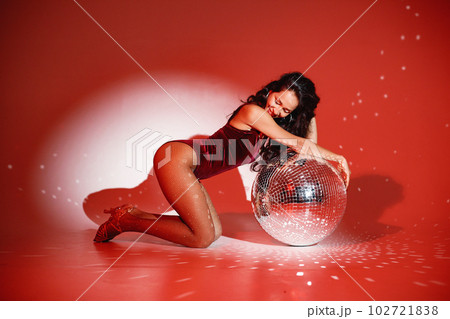Curly brunette girl sitting near big disco ball and posing for a photo. Girl wearing velour bodysuit. Projectors light girl up. Curly brunette girl sitting near big disco ball and posing for a photo. Girl wearing velour bodysuit. Projectors light girl up. 102721838