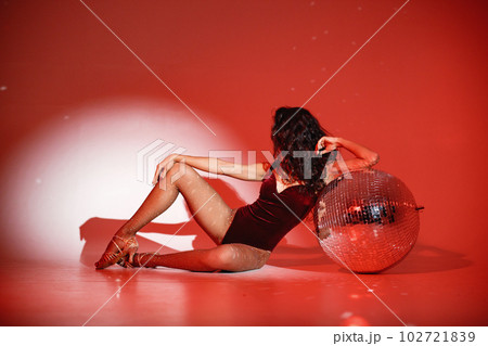 Curly brunette girl sitting near big disco ball and posing for a photo. Girl wearing velour bodysuit. Projectors light girl up. Curly brunette girl sitting near big disco ball and posing for a photo. Girl wearing velour bodysuit. Projectors light girl up. 102721839