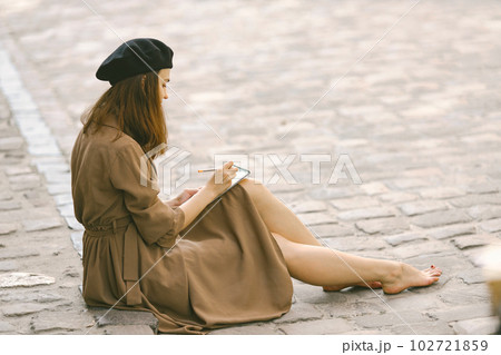 Female painter painting while sitting in a city center. A woman wearing khaki dress and a black hat. Girl drawing a sketch in summer on a street. Female painter painting while sitting in a city center. A woman wearing khaki dress and a black hat. Girl drawing a sketch in summer on a street. 102721859