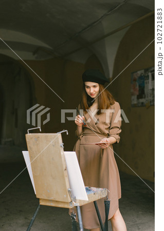 Female painter painting in the city. A woman wearing khaki dress and a black hat. Girl drawing a picture on an easel in summer on a street. Female painter painting in the city. A woman wearing khaki dress and a black hat. Girl drawing a picture on an easel in summer on a street. 102721883