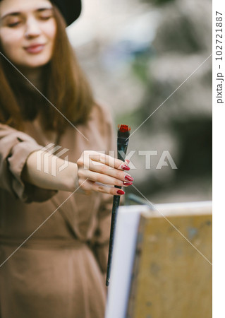Female painter painting in the park. A woman wearing khaki dress and a black hat. Girl measuring the size of an object with a brush. 102721887