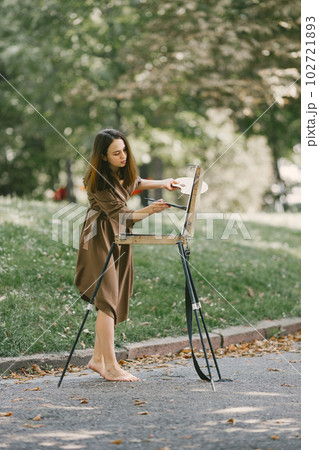 Female painter painting in the park. A woman wearing khaki dress. Girl drawing a picture on an easel in summer on nature. Female painter painting in the park. A woman wearing khaki dress. Girl drawing a picture on an easel in summer on nature. 102721893