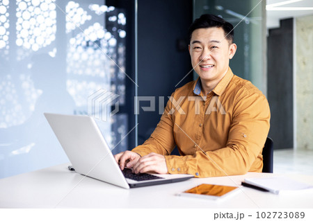 Young and successful asian businessman in working on laptop inside modern office building, happy manager smiling and looking at camera, entrepreneur investor portrait 102723089
