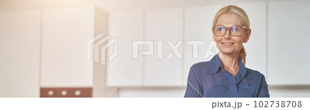 Confident mature businesswoman in glasses smiling aside, working at home using laptop, standing with arms crossed in modern kitchen 102738708