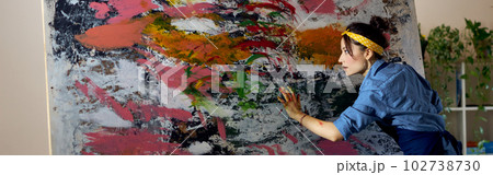Creative young female painter working on a large modern abstract oil painting, applying paint on canvas with fingers in living room at home studio workshop Creative young female painter working on a large modern abstract oil painting, applying paint on canvas with fingers in living room at home studio workshop 102738730