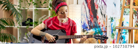 Creative young woman playing guitar while sitting on a bed in modern studio apartment Creative young woman playing guitar while sitting on a bed in modern studio apartment 102738794