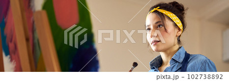 Portrait of young woman in apron holding paintbrush while working on abstract painting at cozy workshop 102738892