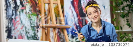 Joyful female painter in apron smiling at camera, holding paintbrush while working on painting, sitting on the floor at home studio workshop 102739074