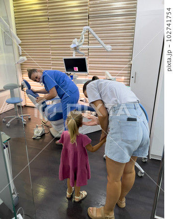 Mom with a little girl stand near the dentist adjusting the dental chair Mom with a little girl stand near the dentist adjusting the dental chair 102741754