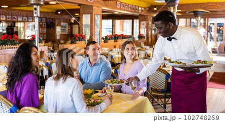 African american waiter serving dishes to cheerful group of adults in restaurant African american waiter serving dishes to cheerful group of adults in restaurant 102748259
