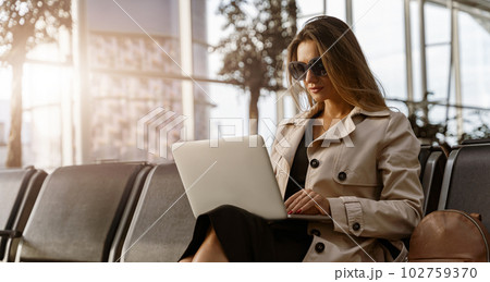 Young businesswoman in sunglasses with luggage in airport waiting room working laptop Young businesswoman in sunglasses with luggage in airport waiting room working laptop 102759370