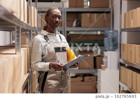 Serious delivery service worker tracking inventory in warehouse portrait. African american shipping business employee checking supply, holding clipboard and writing in checklist Serious delivery service worker tracking inventory in warehouse portrait. African american shipping business employee checking supply, holding clipboard and writing in checklist 102765693