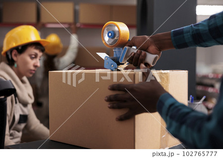 Delivery office manager packing parcel using hand held sticky tape dispenser. African american shipping department worker arms closing cardboard box with adhesive close up 102765777