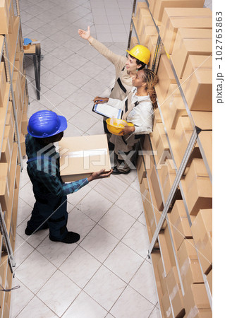 Storehouse employees managing retail stock supply with goods checklist. African american warehouse handler carrying cardboard box and women coworkers checking parcels using orders list top view Storehouse employees managing retail stock supply with goods checklist. African american warehouse handler carrying cardboard box and women coworkers checking parcels using orders list top view 102765863