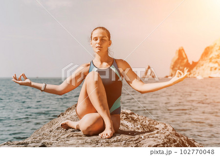 Yoga on the beach. A happy woman meditating in a yoga pose on the beach, surrounded by the ocean and rock mountains, promoting a healthy lifestyle outdoors in nature, and inspiring fitness concept. 102770048