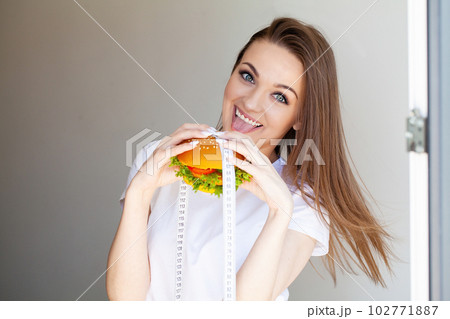 Portrait young woman holding harmful fat burger with white measuring tape. Portrait young woman holding harmful fat burger with white measuring tape. 102771887