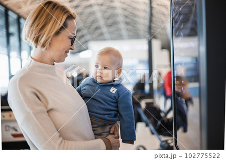 Mother traveling with child, holding his infant baby boy at airport terminal, checking flight schedule, waiting to board a plane. Travel with kids concept 102775522