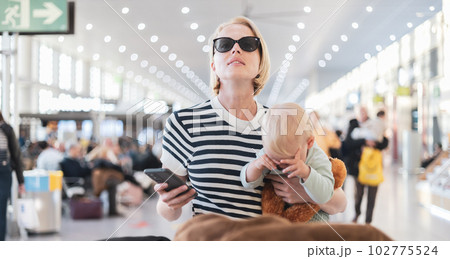 Mother traveling with child, holding his infant baby boy at airport terminal, checking flight schedule, waiting to board a plane. Travel with kids concept 102775524