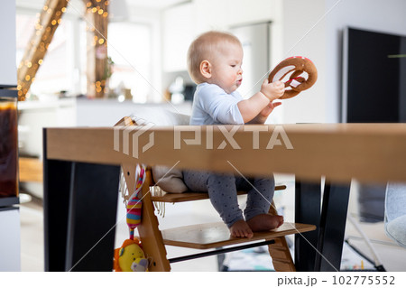 Happy infant sitting at dining table and playing with his toy in traditional scandinavian designer wooden high chair in modern bright atic home. Cute baby playing with toys. Happy infant sitting at dining table and playing with his toy in traditional scandinavian designer wooden high chair in modern bright atic home. Cute baby playing with toys. 102775552
