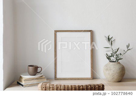 Closeup of picture frame, poster mockup. Vase with olive tree branches on wooden table. Blurred rattan chair. Cup of coffee, books. Summer artistic Mediterranean interior. Working space, home office. Closeup of picture frame, poster mockup. Vase with olive tree branches on wooden table. Blurred rattan chair. Cup of coffee, books. Summer artistic Mediterranean interior. Working space, home office. 102778640