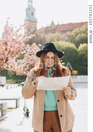Confused traveler woman in hat looking at city map, got lost. Upset young lady backpacker lost in city, checking map in Krakow. Traveling Europe in spring. Navigation, trekking during travelling 102781251