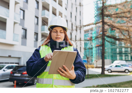 Young architect woman in white hardhat and safety vest using digital tablet outdoors. Female construction engineer. Engineering application 102782277
