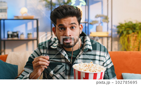 Indian man sits on couch eating popcorn and watching interesting TV serial sport game online at home 102785695