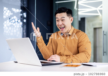 Successful asian man celebrating victory and success received news of good achievement online, businessman looking at laptop screen and holding hand up triumph gesture, worker inside office 102786960