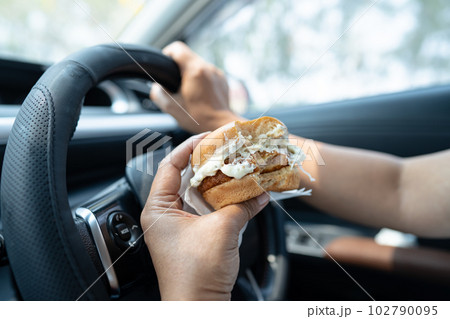 Asian lady holding hamburger to eat in car, dangerous and risk an accident. 102790095