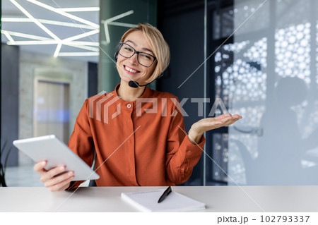 Portrait of a young businesswoman in a headset working in the office on a project, sitting at a table, holding a tablet in her hand, smiling and presenting to the camera. Portrait of a young businesswoman in a headset working in the office on a project, sitting at a table, holding a tablet in her hand, smiling and presenting to the camera. 102793337