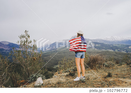 Young woman girl standing on rock cliff with US flag on her back and looking at mountains in background. Female traveller wearing American flag standing mountain top. 4 fourth July Independence Day. Young woman girl standing on rock cliff with US flag on her back and looking at mountains in background. Female traveller wearing American flag standing mountain top. 4 fourth July Independence Day. 102796772