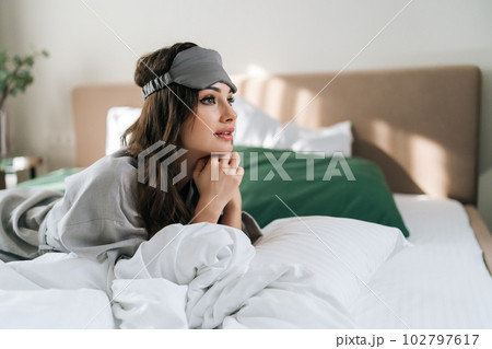 Happy beautiful young woman wearing sleeping mask lying in comfortable bed on blanket in luxury bedroom. Side view of pretty brunette female waking up enjoy good morning ay home. 102797617