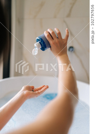 Vertical back view of unrecognizable young woman taking bubble bath, pouring shampoo into hand, enjoying home spa procedure, selective focus. Lovely millennial lady pampering skin, lying in bathtub. 102797656