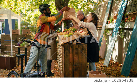 Happy man with bike carrying backpack to deliver food from farmers market stand, picking up box of bio produce. Man courier delivering fruits and vegetables produced by local farmers to customers. 102804239