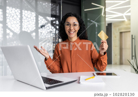 Portrait of happy hispanic woman inside office at workplace, joyful business woman smiling and looking at camera, female worker using laptop at work and holding bank credit card in hands. 102820130