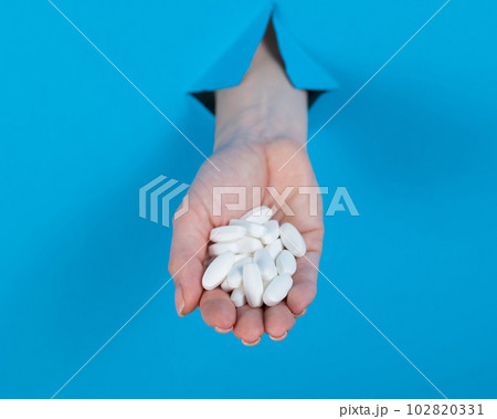 A woman is holding a handful of pills. A woman's hand sticks out of a hole in a cardboard blue background. A woman is holding a handful of pills. A woman's hand sticks out of a hole in a cardboard blue background. 102820331