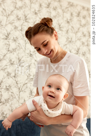 Happy smiling mother holds and hugs her baby in a bright room. Harmonious family, happy motherhood and childhood. 102824635