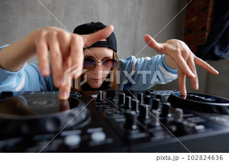 A young attractive DJ girl in a black baseball cap, blue sweater and sunglasses plays music at the DJ mixing board. Musical creativity and energy. 102824636