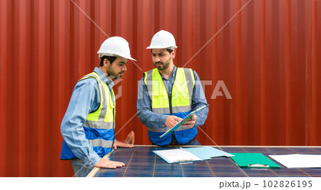 Shipment worker pointing at shipping schedule on clipboard, explain to colleague about planning for next shipment. They use solar cell panel as a table. A large container is in the background Shipment worker pointing at shipping schedule on clipboard, explain to colleague about planning for next shipment. They use solar cell panel as a table. A large container is in the background 102826195
