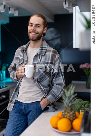 Man with beard holding cup with coffee, standing by kitchen counter with modern light interior. Man with beard holding cup with coffee, standing by kitchen counter with modern light interior. 102829487