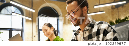 Young smart guy wearing eyeglasses working on laptop while sitting at desk with his female colleague in the modern coworking space or office Young smart guy wearing eyeglasses working on laptop while sitting at desk with his female colleague in the modern coworking space or office 102829677