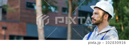 Young bearded male engineer in hard hat looking away, posing outdoors while working on cottage construction 102830916