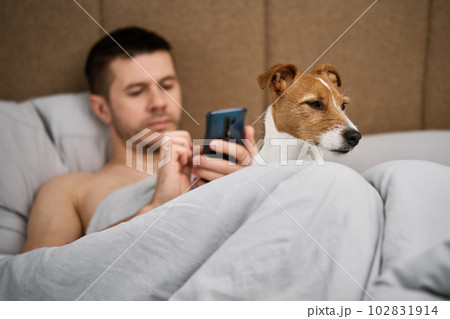 Man with cute dog chilling in bed at morning and use smartphone, lazy morning. Spending time together, pet affection 102831914