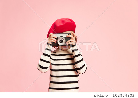 Portrait of little girl, cild in striped sweater and red beret taking photo with vintage photo camra agaisnt pink studio background 102834803