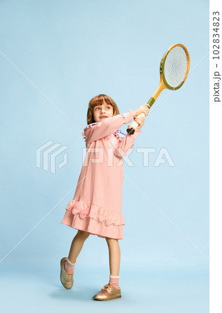 Full-length portrait of little girl in pink dress posing with tennis racket, playing against blue studio background Full-length portrait of little girl in pink dress posing with tennis racket, playing against blue studio background 102834823